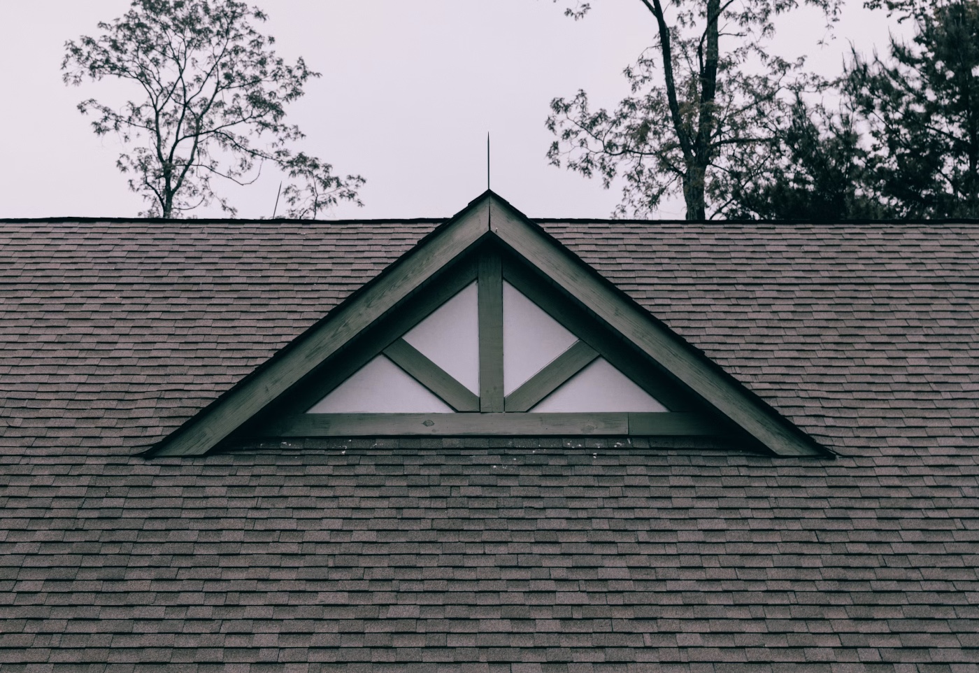 Peaked roof gable with triangular window and spire against gray sky and trees