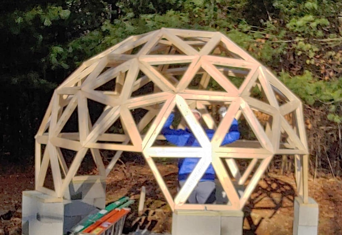 Wooden geodesic dome structure under construction with person inside wearing blue shirt